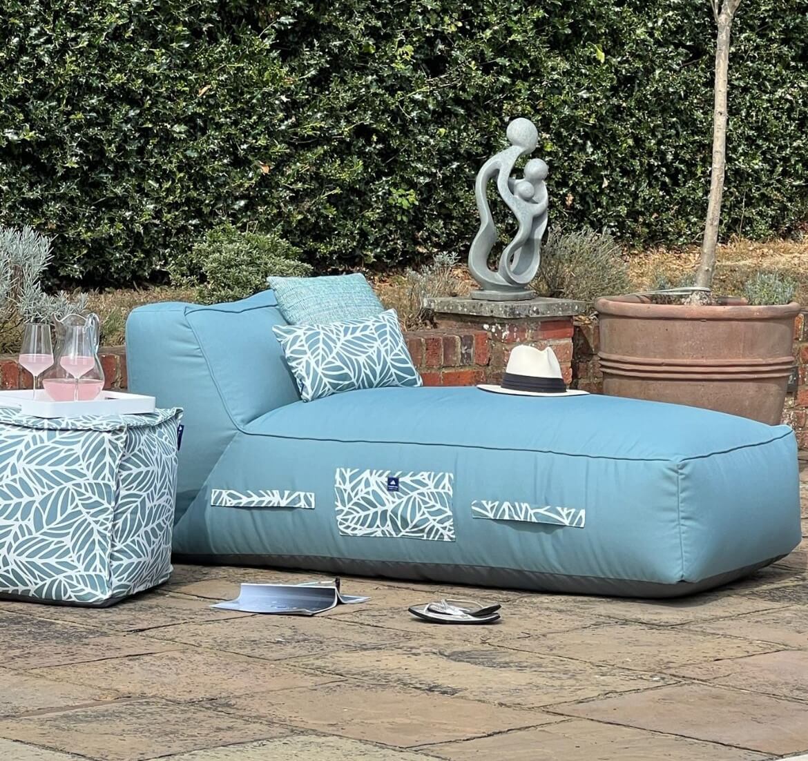 Ocean blue fabric cushioned sun lounger with palm-patterned outdoor cushions. There is a jug of cool pink lemonade and two glasses on a palm-patterned pouffe.