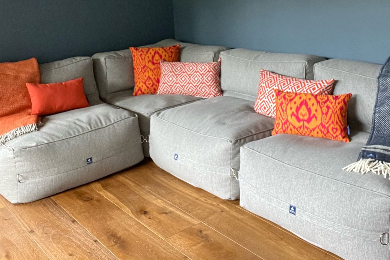 A large L-shaped cushiony sofa in light grey in the corner of a room. The sofa is covered with orange and paprika patterned cushions