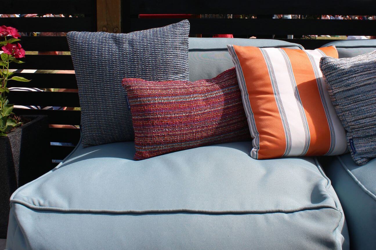 A photograph of four coloured and patterned outdoor throw cushions on a blue fabric garden sofa. The cushions are orange and white striped, a textured deep red, and a woven blue and aquamarine