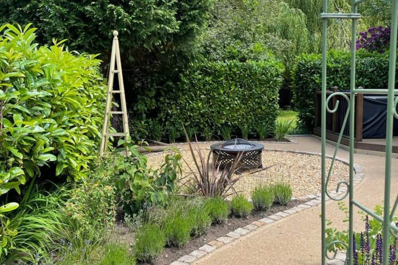 A gravel path lined with brick pavers curls round a corner and opens up into a circular patio with a firepit at its centre. Lush greenery and plant obelisks surround both patio and path.