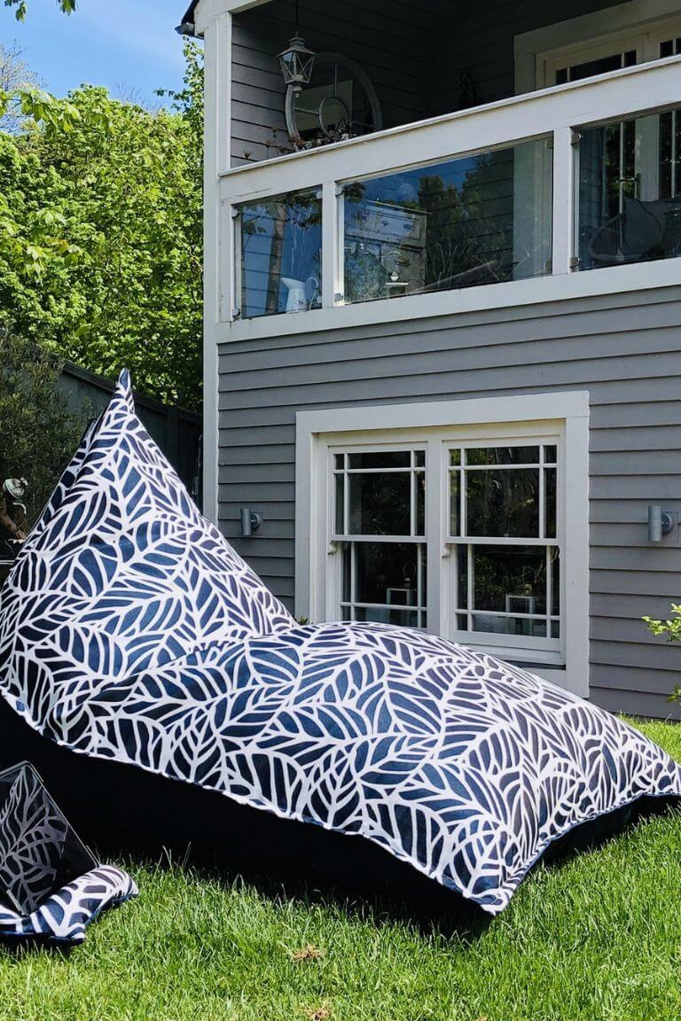 A photograph of a large outdoor bean bag lounger sat on grass in the sunshine. The fabric of the garden lounger is navy and white in a palm-leaf pattern.