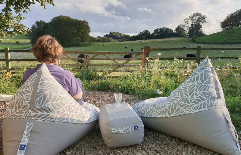 Two bean bag chairs sit either side of a low snack table on a gravel patio. A woman sits comfortable on one of the outdoor bean bag chairs, enjoying a beautiful view.