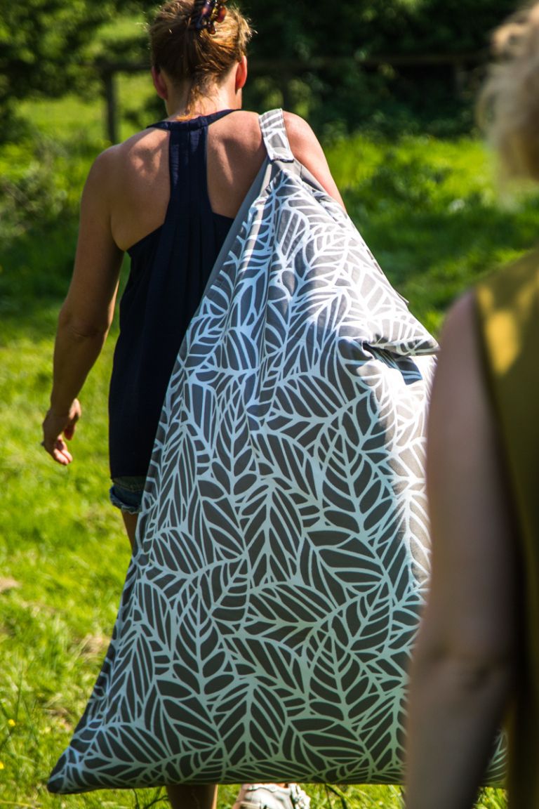 A photograph of a woman walking carrying an outdoor bean bag over her shoulder by a handle strap sewn into its side. The fabric of the bean bag is grey and white in a palm leaf pattern.