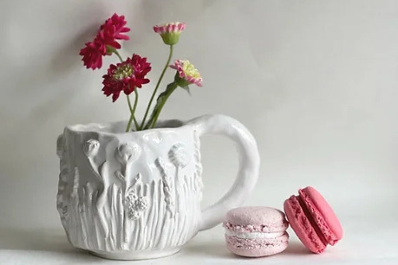 A pure white large china mug decorated liberally with flowers and grasses. Real pink flowers have been put in the mug and two pink macarons lay next to it.