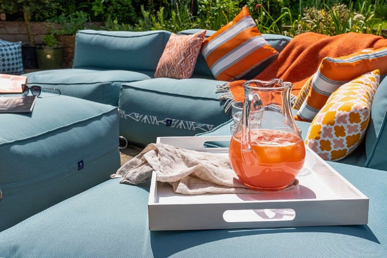 A soft blue cushioned outdoor sofa is placed in the sunshine and covered with orange and white throw cushions as an invitation for conversation and socialising.