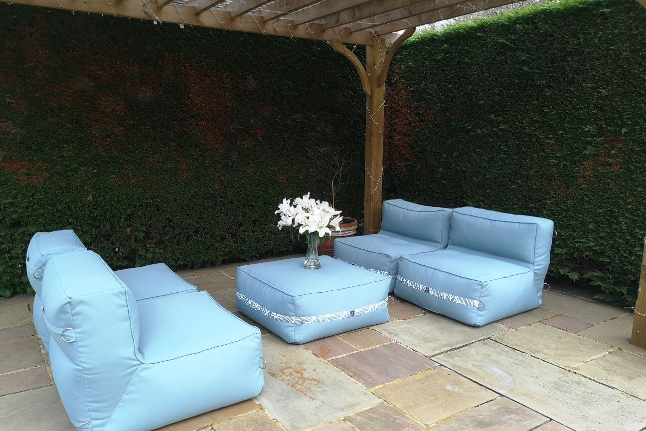 A light blue sectional garden sofa is arranged with two pairs of soft chairs facing each other across an ottoman table ready for alfresco dining.