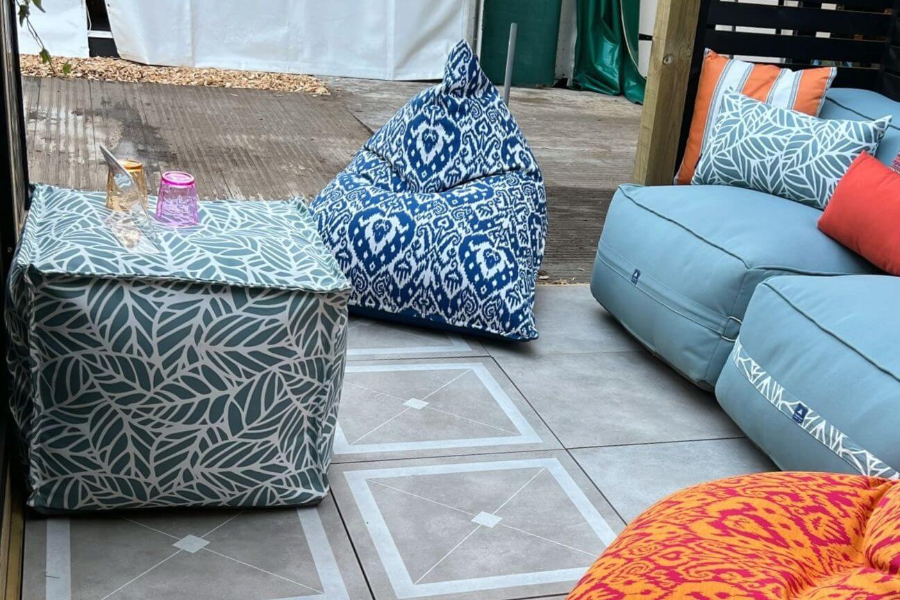 A phhotograph of a small garden furnished with outdoor furniture including a modular garden sofa, two garden bean bags and a waterproof pouffe being used as a table. There are two glasses sat on the outdoor pouffe.