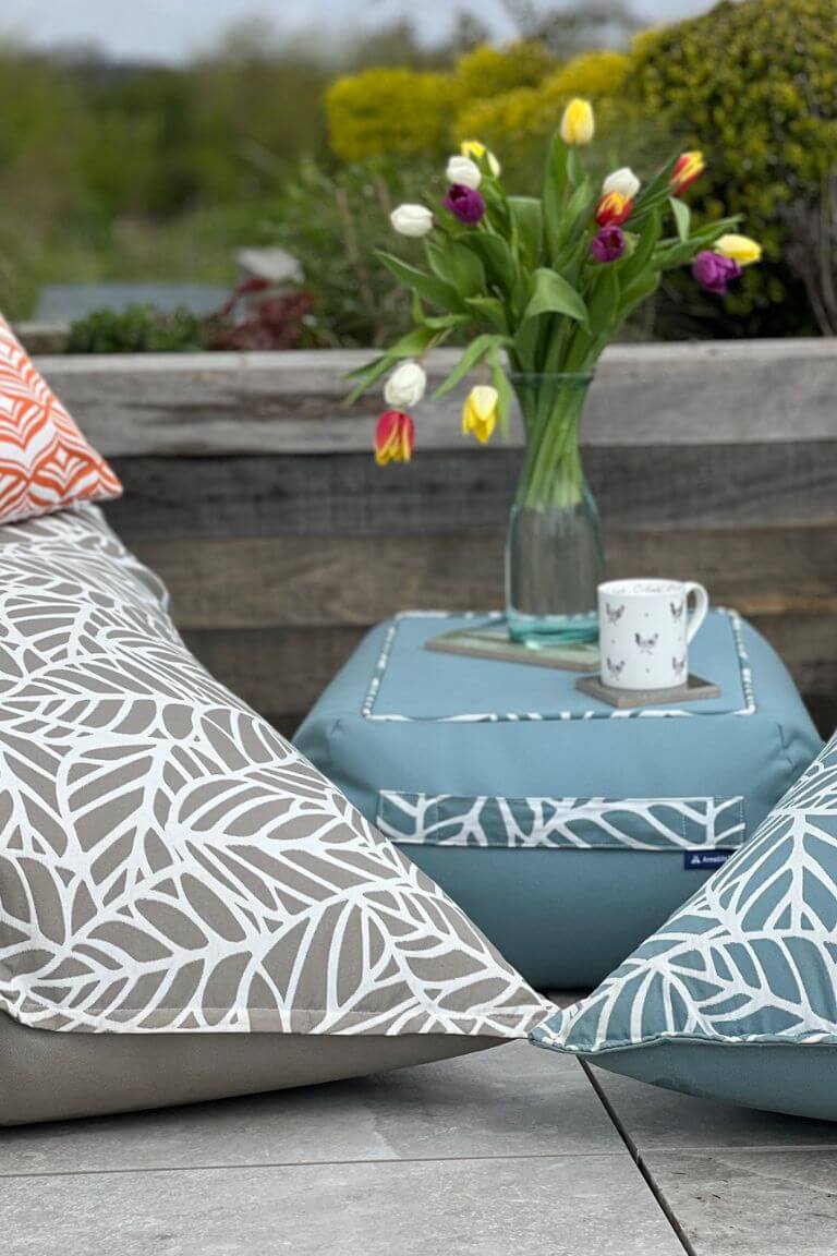 A photograph of a low, small, rectangular bean bag coffee table. A cup of tea and vase of flowers are sat on the hard top of the table. The table is ocean blue with trim in a palm-leaf pattern.