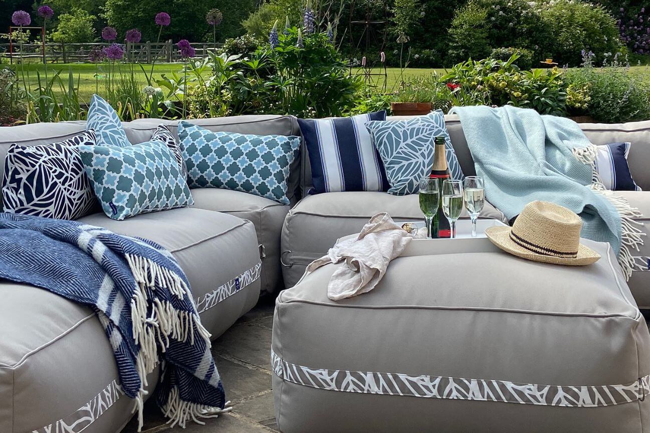 A soft garden sofa made from fabric is on a patio surrounded by greenery and purple flowers. The sofa is covered with striped and patterned garden cushions. A bottle of champagne is waiting to be poured.