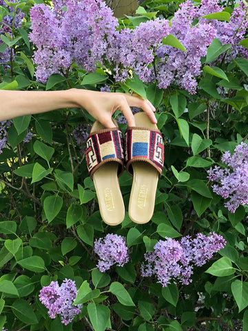 womens kilim sandals in sumak style being held in front of green plant with purple flowers. 