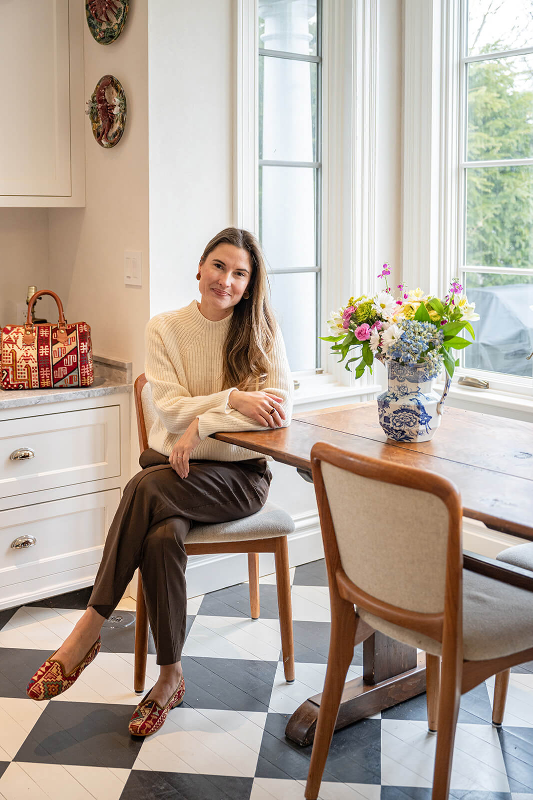 Cordelia Fasoldt, a white woman with dark hair, seated at her kitchen table, wearing Artemis Design Co. Sumak Loafers.  There are french doors to outside to her left, and checkerboard stone floors.