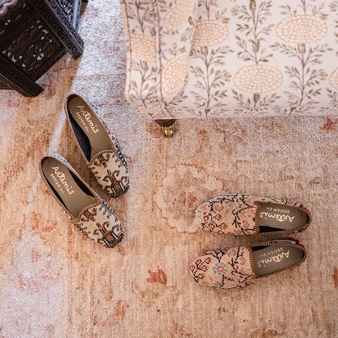 Looking down at a soft pink patterned rug, with the corner of an ottoman visible in the top right section.  There are two pairs of light, neutral colored Oriental Carpet shoes on the floor: a cream and brown pair of loafers at left, and a pale pink pair of Smoking Shoes at right.