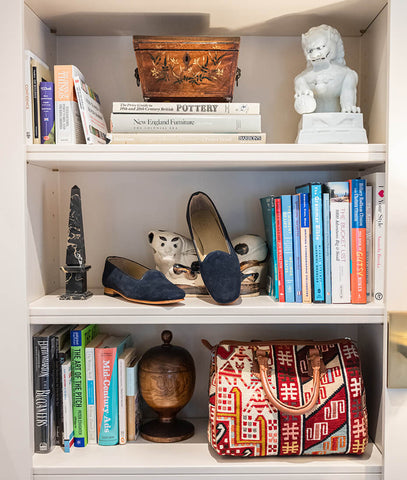 White shelves with books and objects displayed on them, as well as a pair of navy suede women's loafers on the middle shelf, and a Sumak Baby Duffle handbag on the bottom shelf.