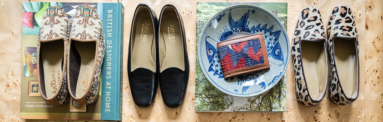 A row of items on a burled wood coffee table: from left, neutral Women's Oriental Carpet Loafers; Black Leather Loafers; a Kilim Wallet in a blue & white ceramic bowl; Leopard patterned Pony Hair Loafers.