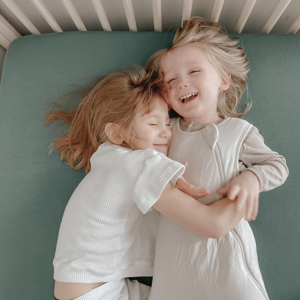 two toddlers laying in twin bed one is wearing a white shirt one is wearing a sleep bag