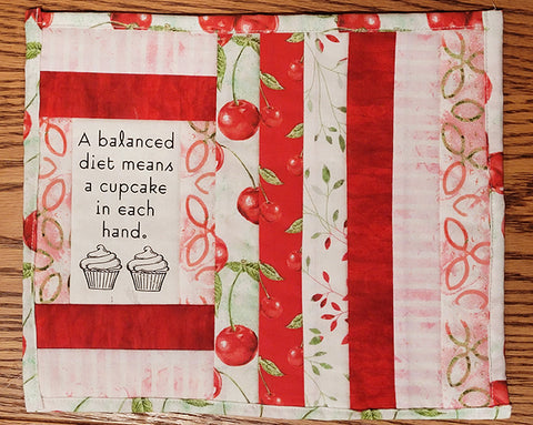 Image shows a small red, green and white quilt with 2 cupcakes that says, "A balanced diet means a cupcake in each hand."