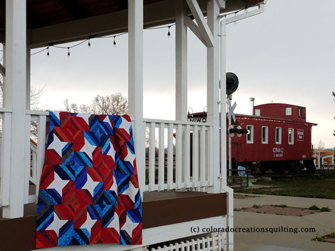 Red, white and blue quilt hangs over the railing of a white gazebo with a red caboose in the background.