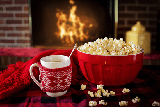 Mug of hot cocoa and a red bowl of popcorn in front of a fire place