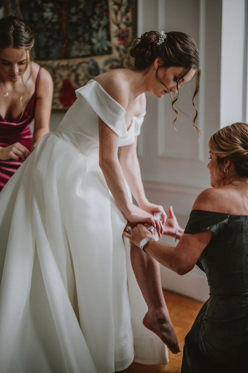 bride getting ready on wedding morning with mom's help putting on her garter