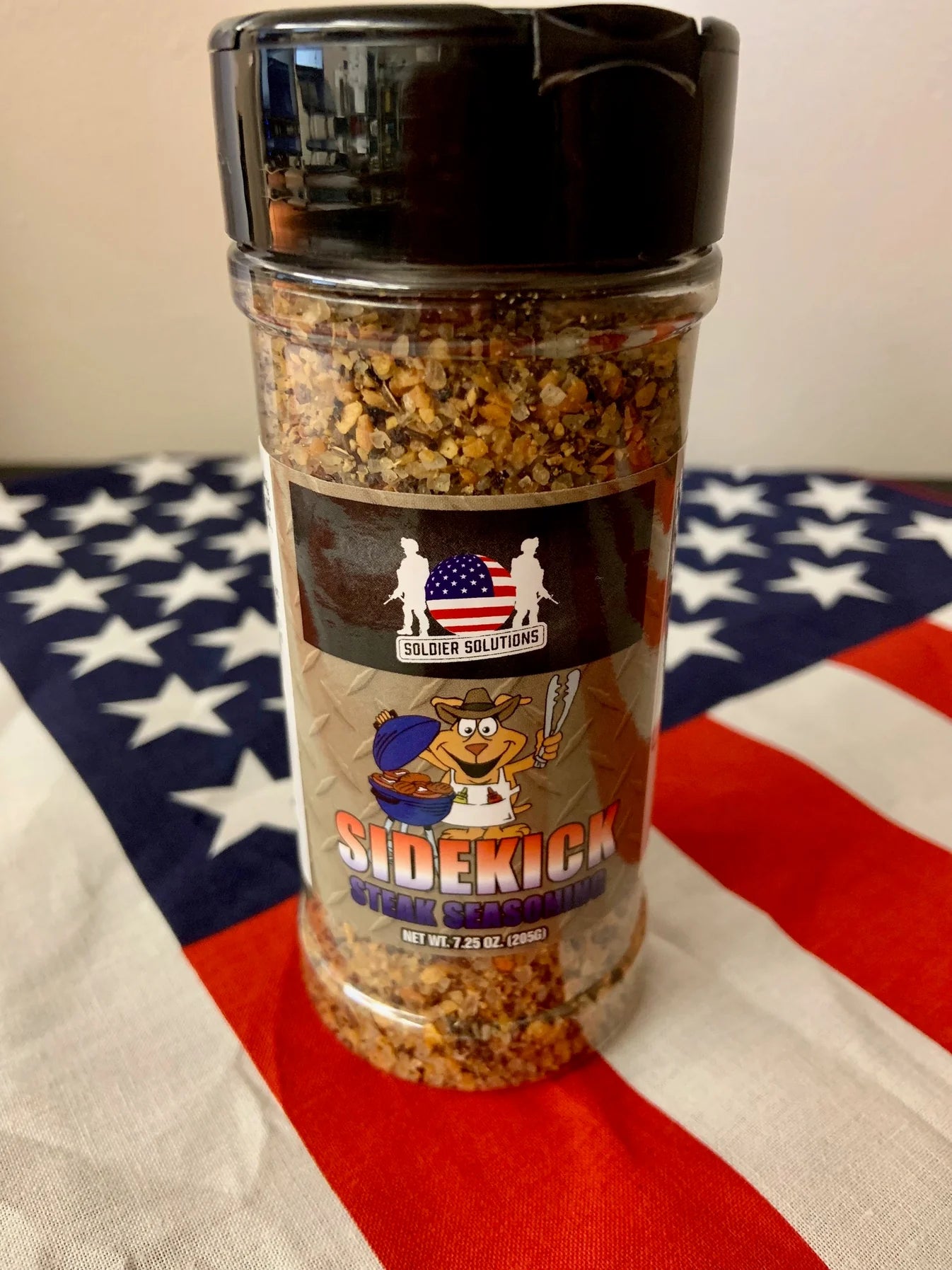 Close-up of Soldier Solutions' Sidekick Steak Seasoning bottle placed on an American flag background—perfect patriotic gift for dads who love grilling and flavor.