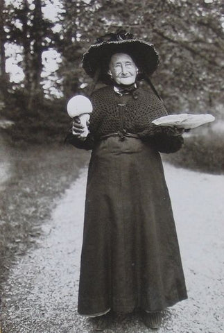 A black and white photograph of an older woman in a black dress with a hat fashioned as a witch hat. She stands in the middle of a road holding two large mushrooms one in each hand.