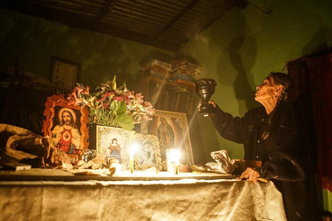 An eldery woman on the right side of a table, raising a gourd shaped container. On the table are candles, flowers, and depictions of Jesus Christ.