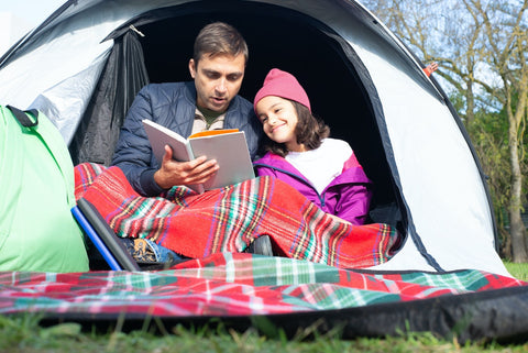 A father reading to his daughter on a camping trip.