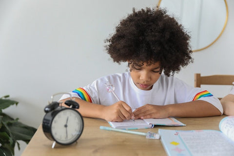 Boy sitting at a desk and doing his homework with an alarm clock next to him