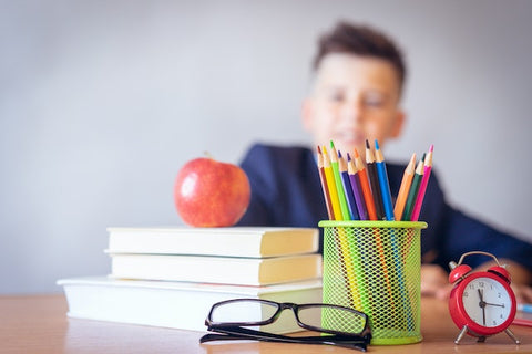 Apple on top of a pile of books, next to a red alarm clock and pair of glasses on a desk where a little boy is sitting