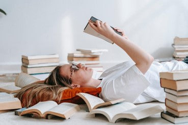 A woman lying on the floor reading a book while being surrounded by many books.