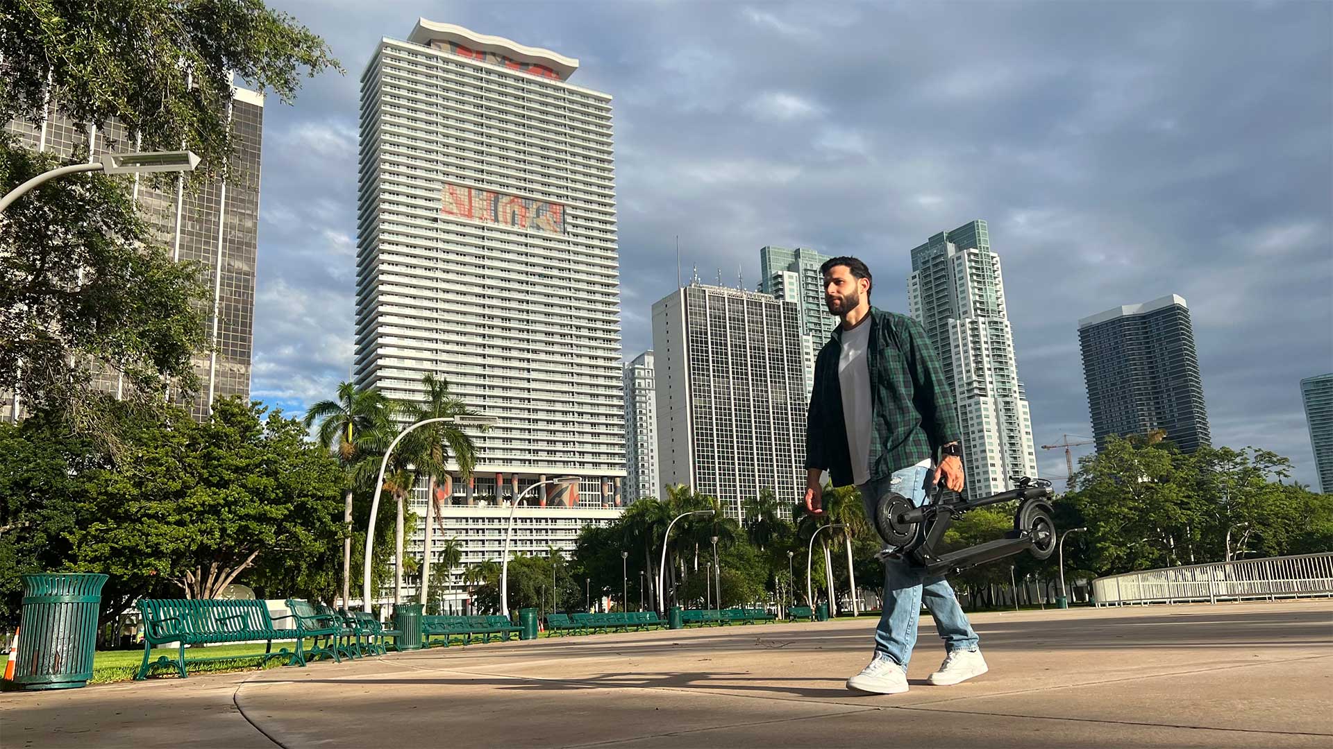 Man carrying an electric scooter in a modern city park with skyscrapers in the background, representing the urban landscape covered in the Complete US Electric Scooters Laws Guide for 2024.