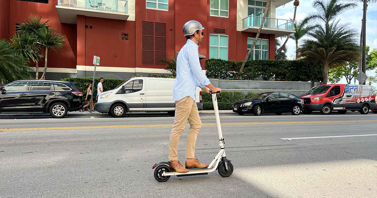 A man in casual business attire rides a white electric scooter on a city street, showcasing an efficient and green transportation option.