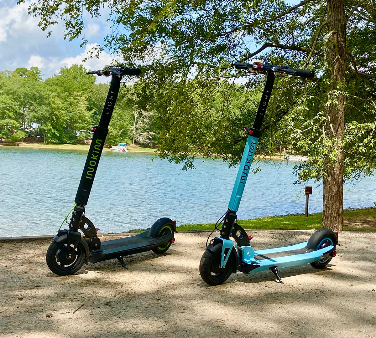 Two electric scooters, one black and one teal, rest side by side on a sandy lakeside path, offering a sustainable way to enjoy nature.