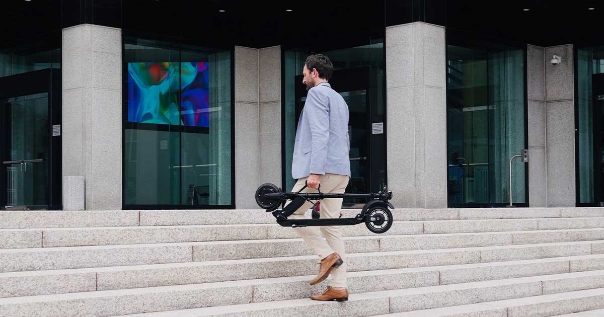 A professional carries a folded electric scooter up the steps of an office building, symbolizing the integration of personal electric vehicles into daily work life and the shift from recreational gadget to transport essential.