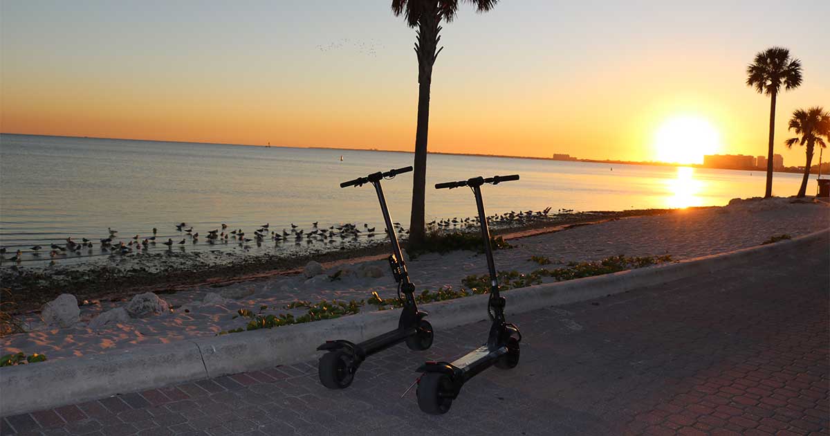 Two electric scooters stand against a scenic sunset background by the ocean, highlighting eco-friendly transportation options.