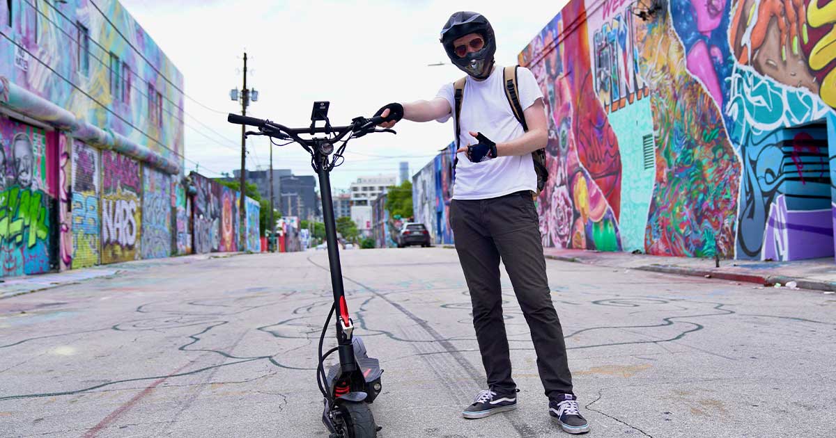 A trendy rider with a helmet giving a thumbs-up while standing next to his electric scooter in an urban alley adorned with colorful graffiti, reflecting the youthful energy driving the electric scooter movement.