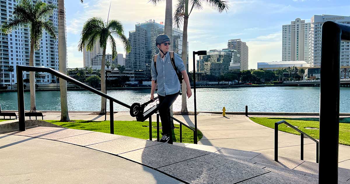 A person carries an electric scooter up the stairs by a sunlit waterfront, symbolizing the versatility and contribution of electric scooters to reducing carbon emissions in coastal cities.