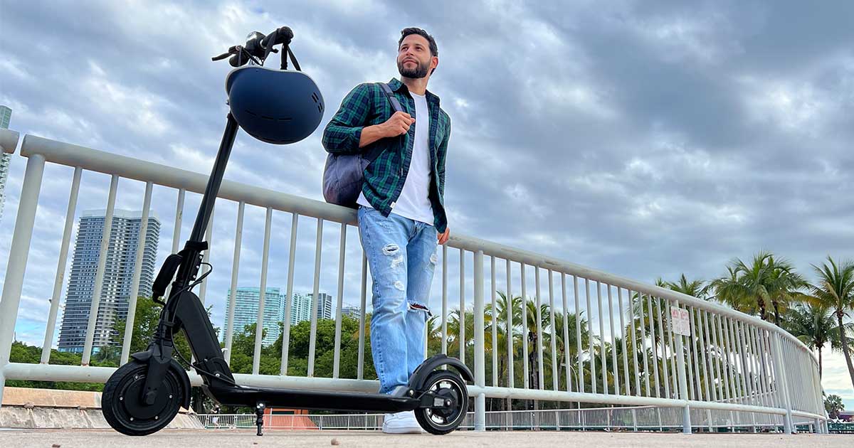 A man standing next to an electric scooter with a helmet hanging on the handlebar, signifying preparedness for road safety while sharing streets with scooters.