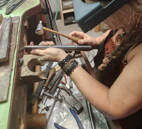 Color photo of a woman working at a wooden bench fashioning a ring with traditional tools