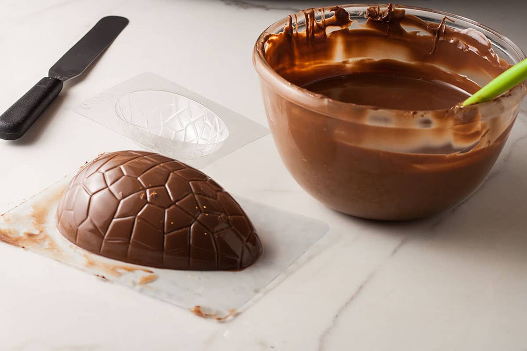 Moule à œufs de Pâques en chocolat enduit, à l'envers sur le plan de travail, avec un bol de chocolat tempéré sur le côté et un moule inutilisé derrière.