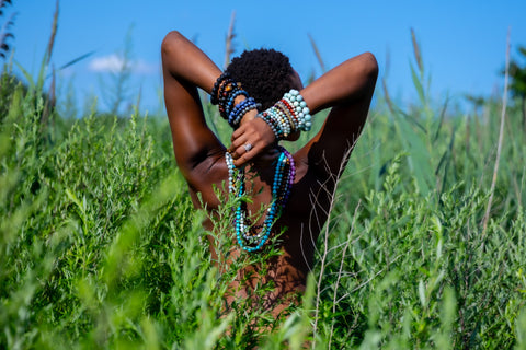 Woman standing in tall grass nude wearing gemstone jewelry.