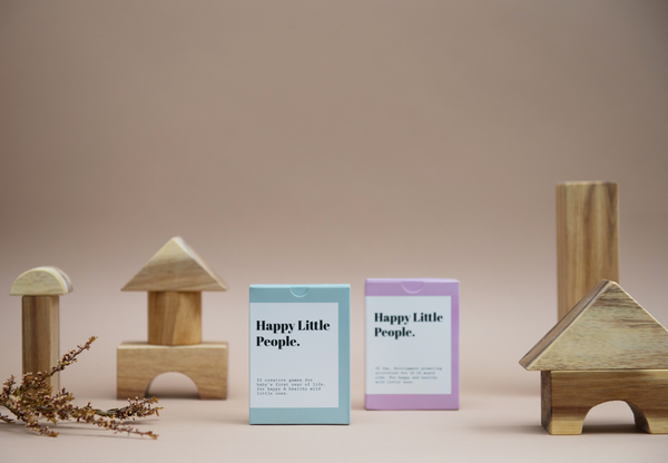 In the foreground, there is one blue deck of cards and one purple, surrounded by dried flowers and wooden blocks. Happy Little People in a serif font is printed on the front on a white label.