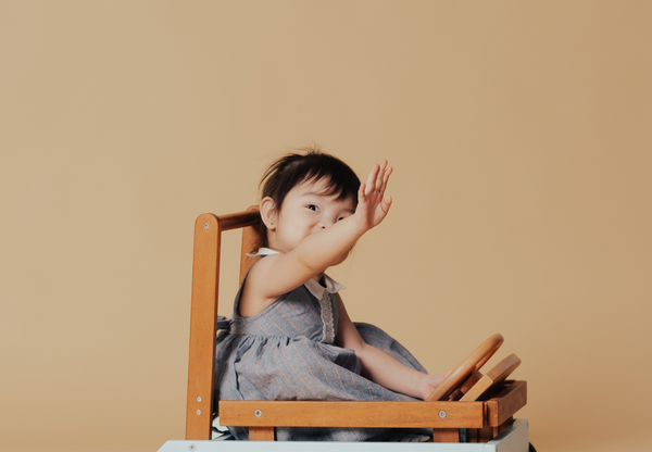 A two-year-old girl waves off-camera with one hand while the other rests on a wooden steering wheel. She is wearing a gray pinstripe dress with a white color against a taupe background.