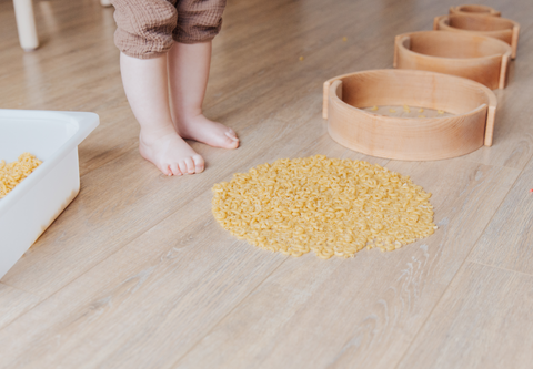 Two bare feet clothed in brown pant legs stand before several wooden rings atop a beige floor. There is a circle of dried macaroni noodles and a white bin.