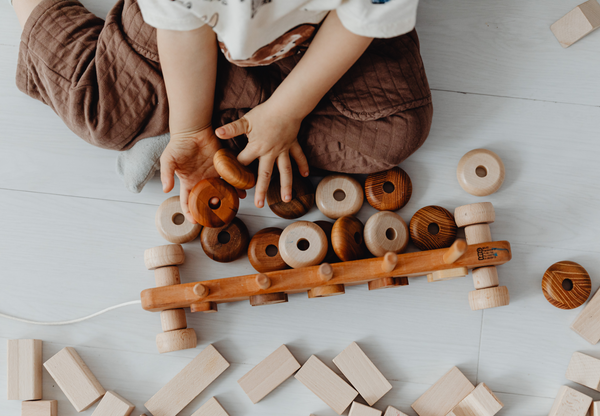 A child in brown pants and a white shirt sits on a white wood floor surrounded by tan wooden blocks. The child is playing with wooden rings and a stacking pull toy.