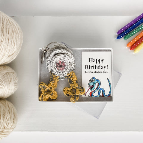 white chicken butt keychain with an illustrated snake art card on a white box with a clear lid