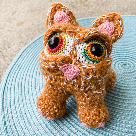 Crocheted orange cat with big rainbow eyes on a sky blue mat