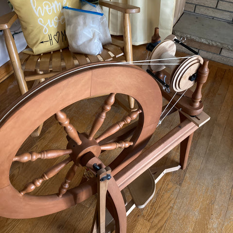 photo of a wooden spinning wheel in front of a rocking chair with a yellow pillow and one of the plastic bags of dog fur