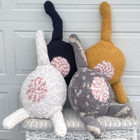 set of 4 crocheted cat butt pillows on an old wicker table in black, orange, white, and gray tabby