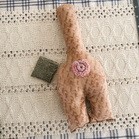 Orange fabric cat butt kicker with a crocheted butthole sitting on a lace table runner next to a small bag of homegrown catnip